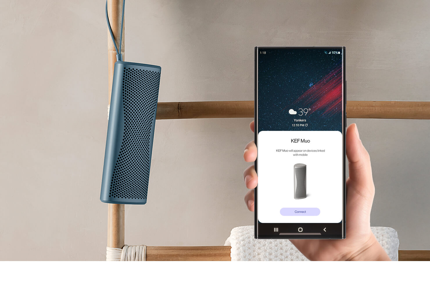 Blue Muo Bluetooth speaker hanging from carrying strap in bathroom is being controlled via phone.