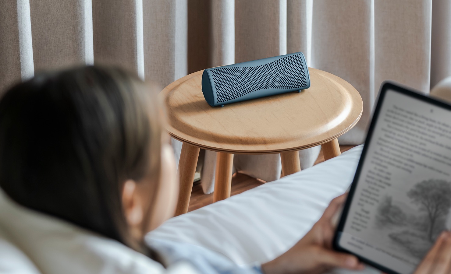 Muo Bluetooth-luidspreker op een houten bijzettafeltje naast iemand die op een tablet zit te lezen.
