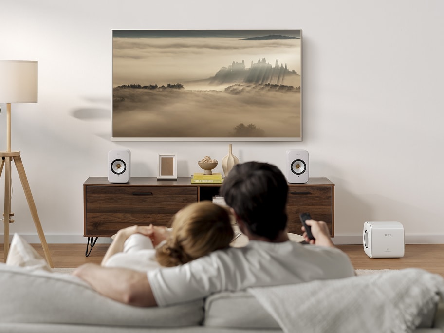 Couple in living room watching TV connected to LSX II speakers in cotton white and KC subwoofer.