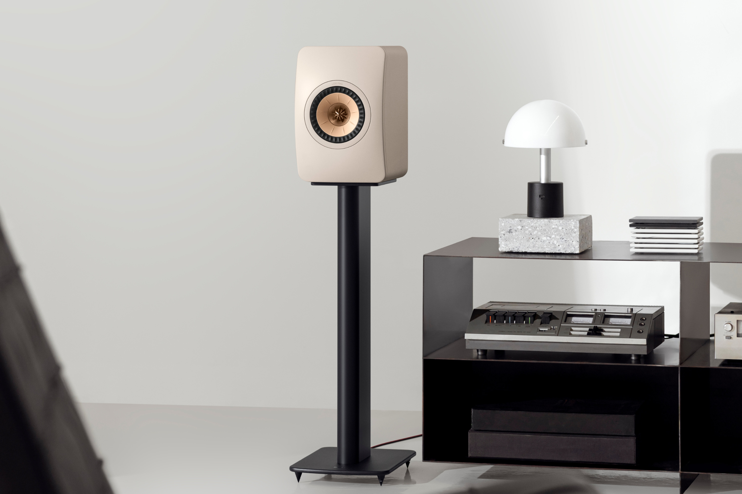 Modern beige bookshelf speaker on a black floor stand in a minimalist living room, beside a black shelving unit with a retro audio system and decorative lamp