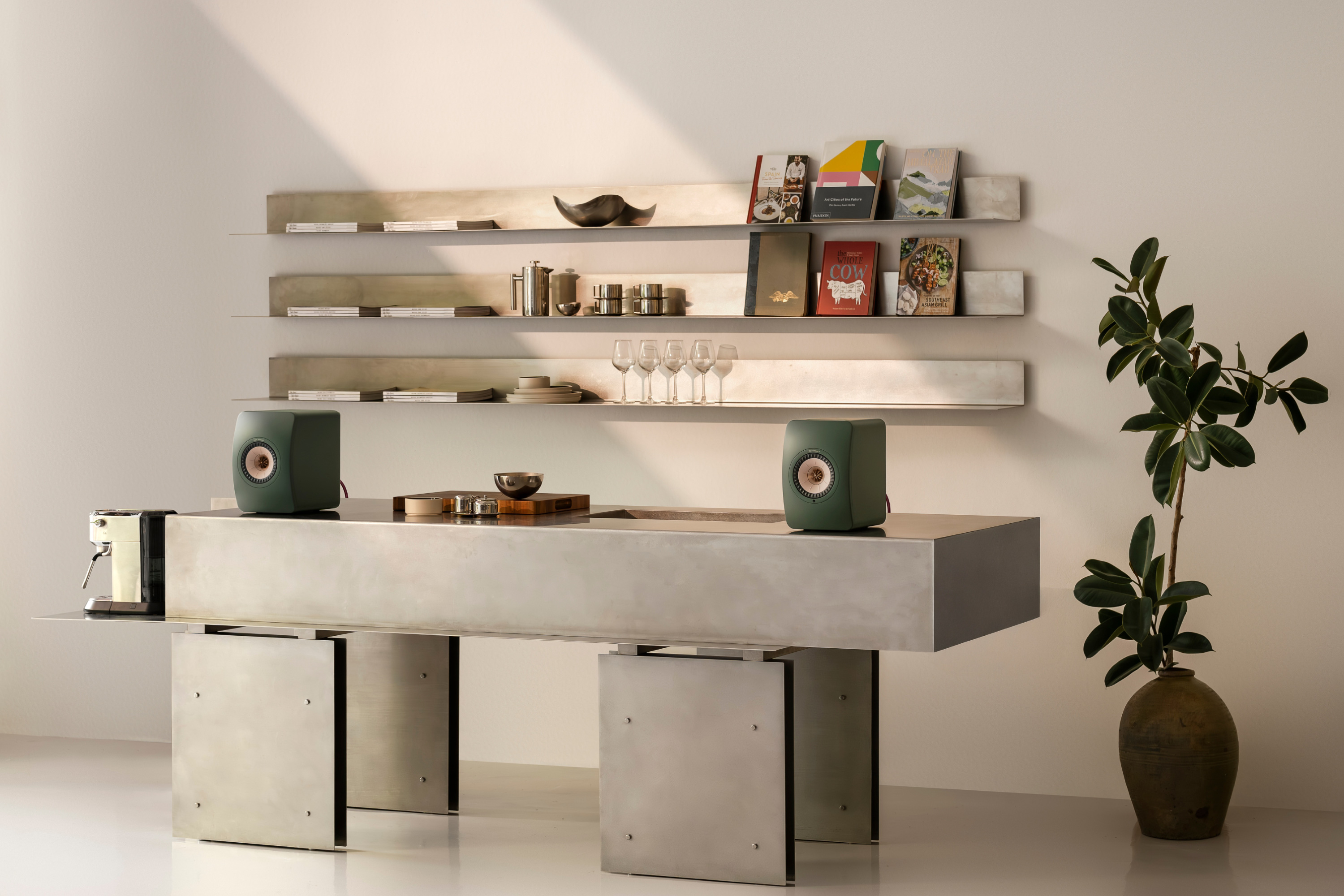 Pair of green bookshelf speakers on a modern concrete table in a minimalist kitchen with open shelving, cookbooks, glassware, and a potted plant.