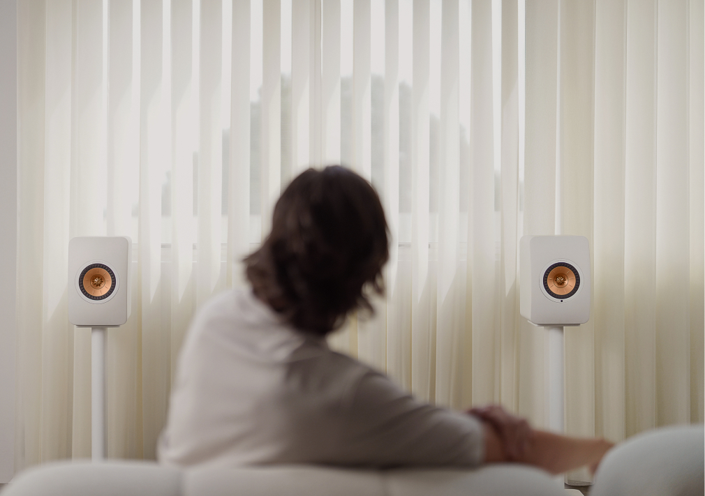 Una persona sentada en un sofá, con un par de altavoces inalámbricos LS50 Wireless II blancos con drivers de cobre colocados sobre soportes delante de unas grandes persianas verticales.