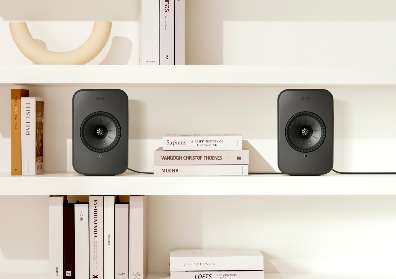 Deux enceintes bibliothèque KEF noires posées sur une étagère blanche, entourées de livres soigneusement disposés et d’objets décoratifs, dans un salon moderne.