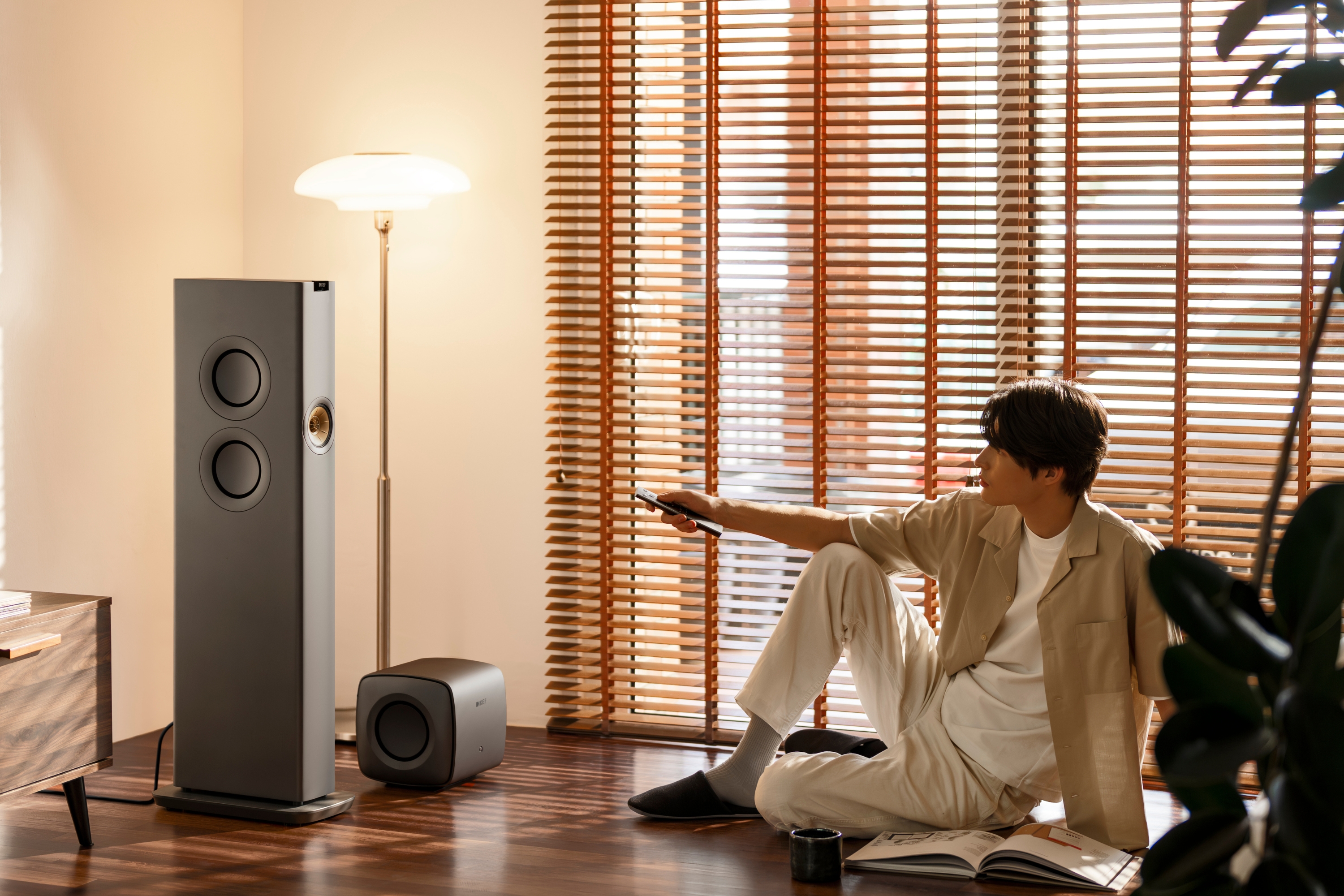 Un homme assis sur un sol en bois dans une pièce chaleureuse et baignée de soleil utilise une télécommande pour contrôler une paire d’enceintes colonne KEF et un caisson de basses compact, entouré de stores en bois, d’un lampadaire moderne et d’un magazine.