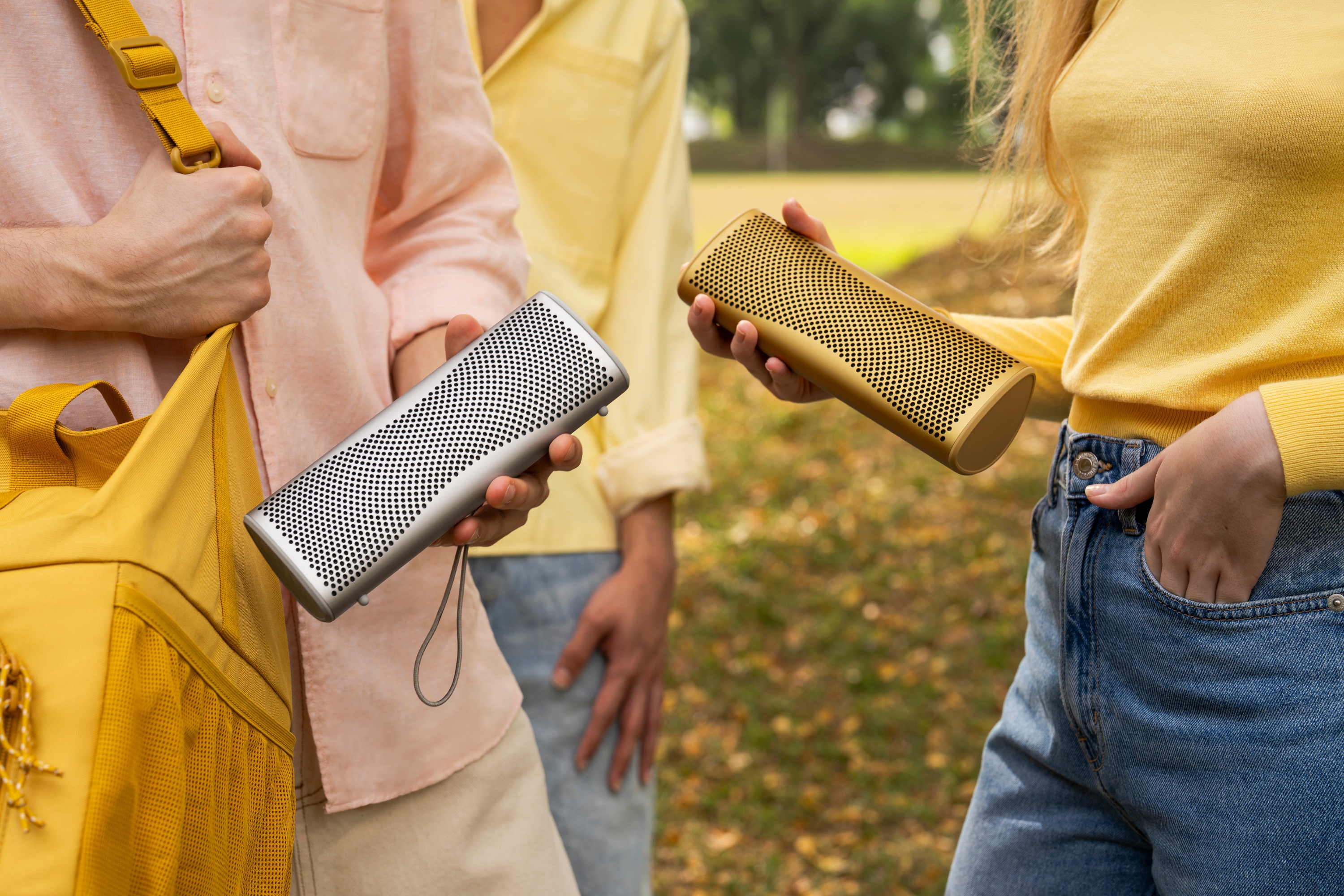 Nahaufnahme von zwei Personen, die kompakte tragbare Lautsprecher in Silber und Gelb im Freien halten.