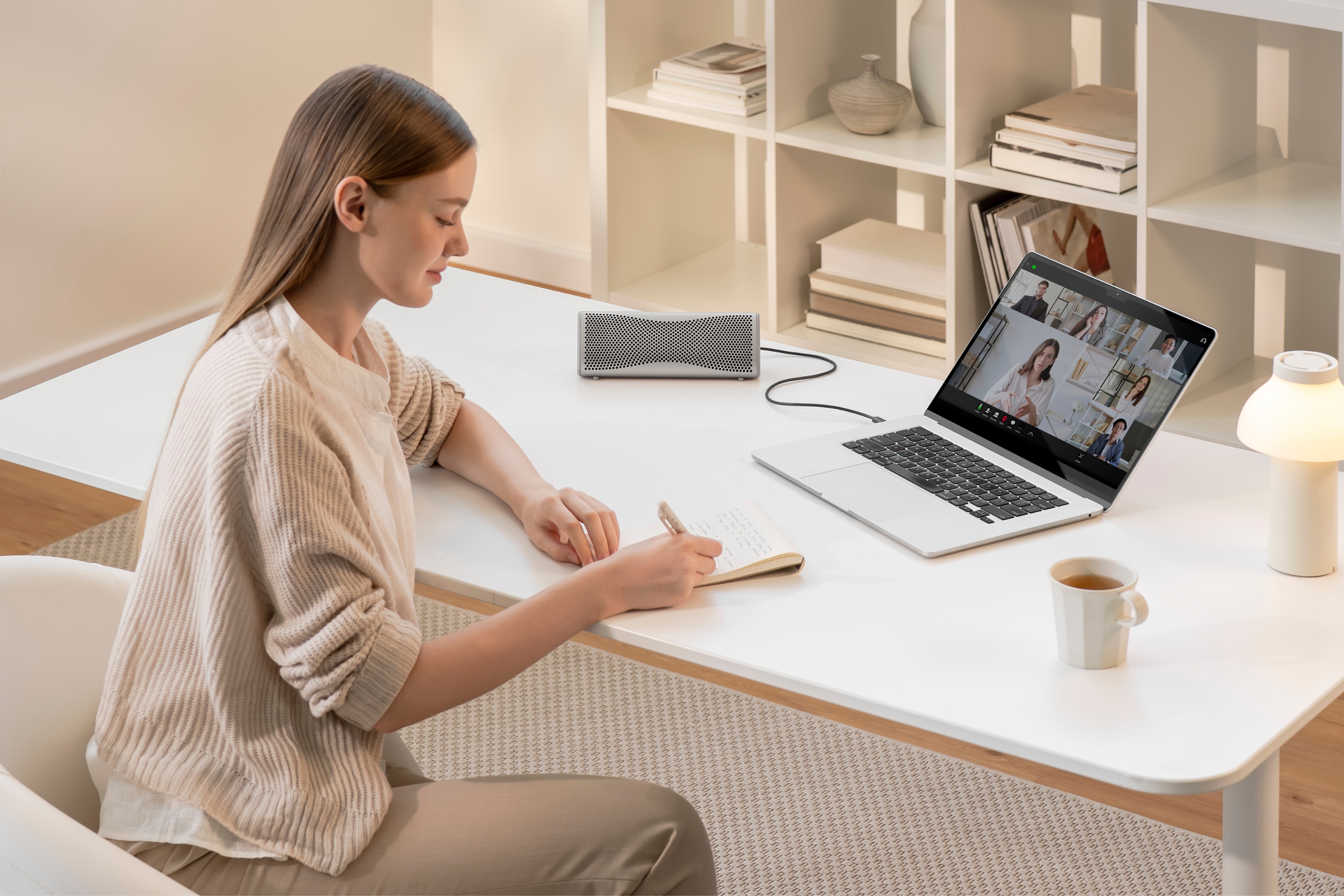 Person macht sich Notizen an einem Schreibtisch während eines Videoanrufs, ein kompakter Lautsprecher ist mit einem Laptop verbunden.