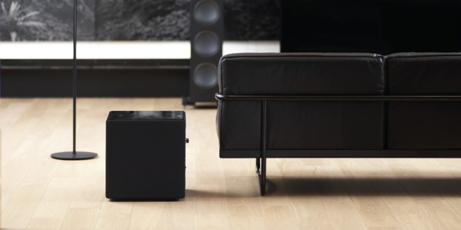 Système HiFi KEF composé d’une enceinte colonne noire et d’un caisson de basses, installé dans un salon élégant avec parquet, canapé en cuir noir et une grande enceinte en arrière-plan.