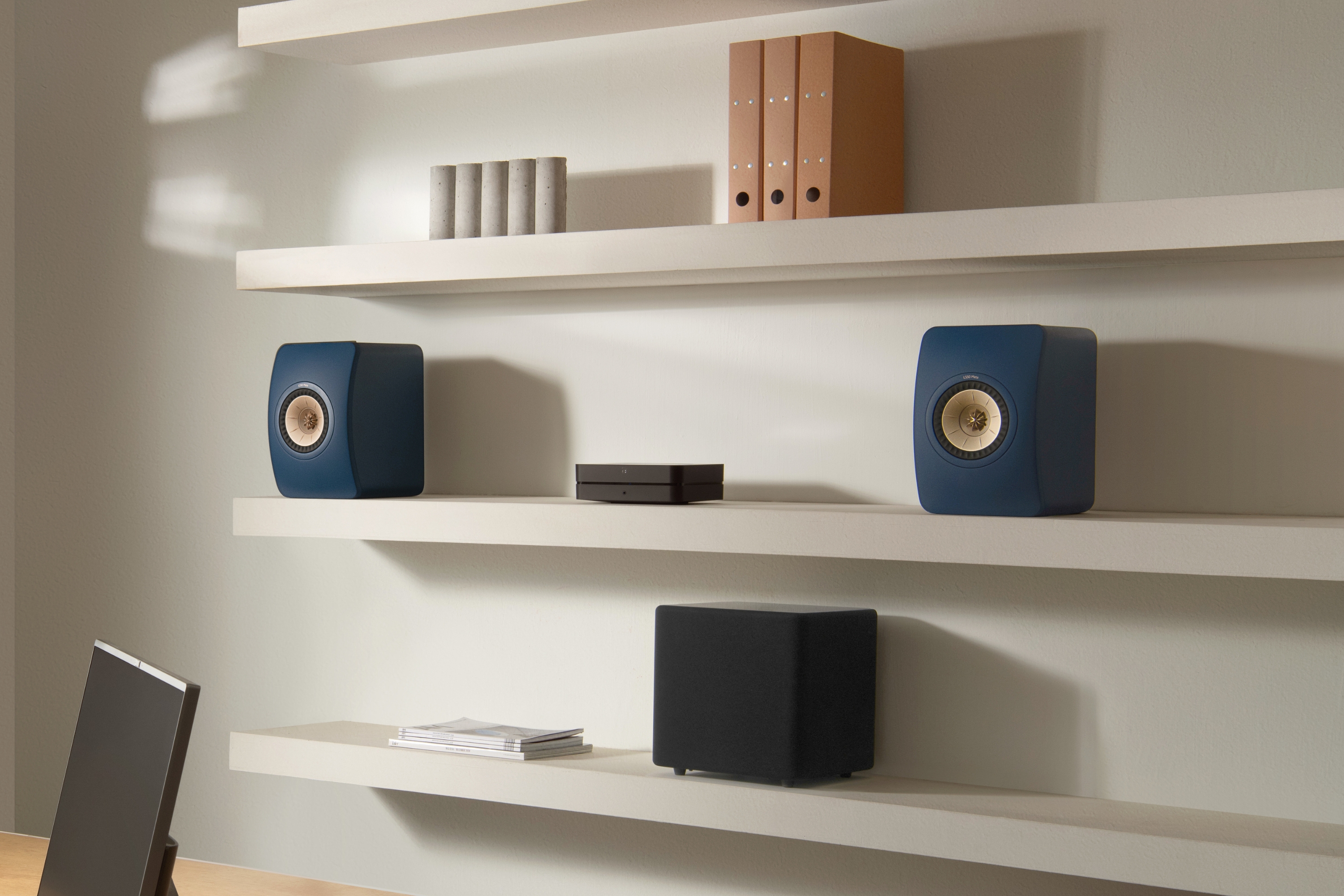 Enceintes bibliothèques KEF bleues audacieuses sur des étagères murales flottantes dans un bureau à domicile moderne, accompagnées d’un caisson de basses et d’accessoires de rangement.