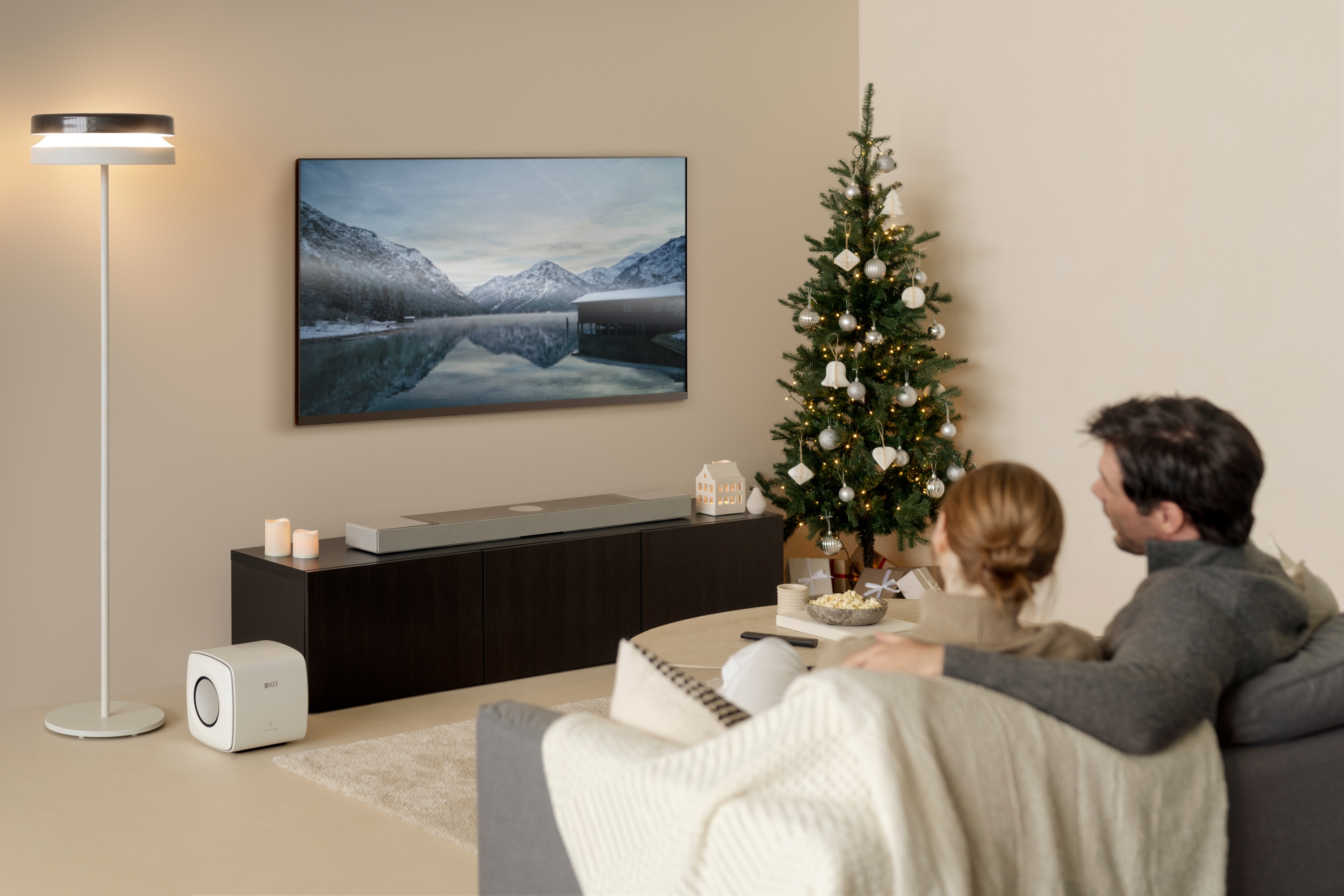 Een koppel ontspant op de bank en kijkt televisie naast een kerstboom, met een stijlvolle zilverkleurige soundbar en een witte subwoofer voor een echte thuisbioscoopervaring.