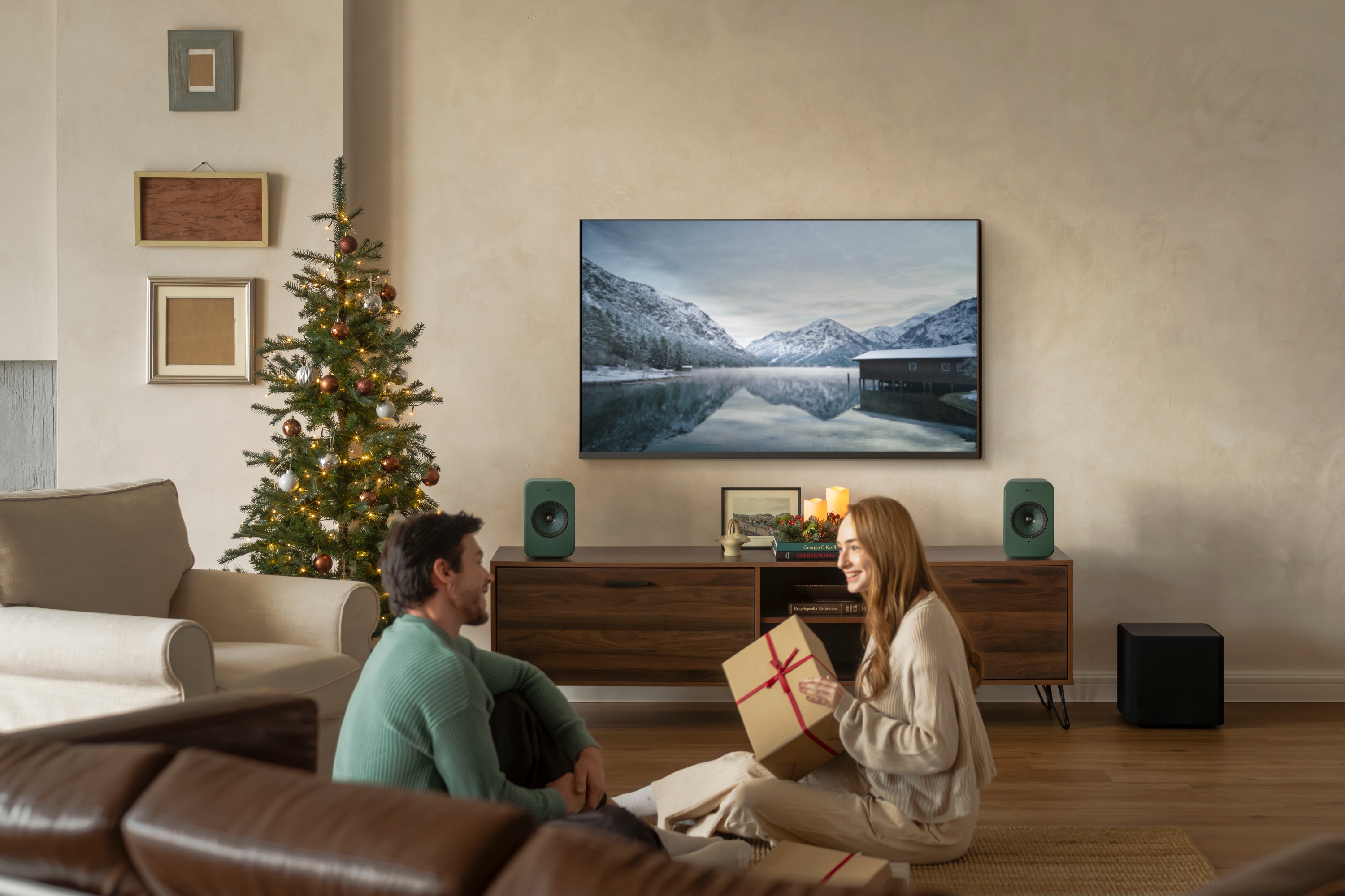 Een koppel zit bij de kerstboom en wisselt cadeaus uit in een gezellige woonkamer met turquoise draadloze speakers en een televisie met een winters landschap.