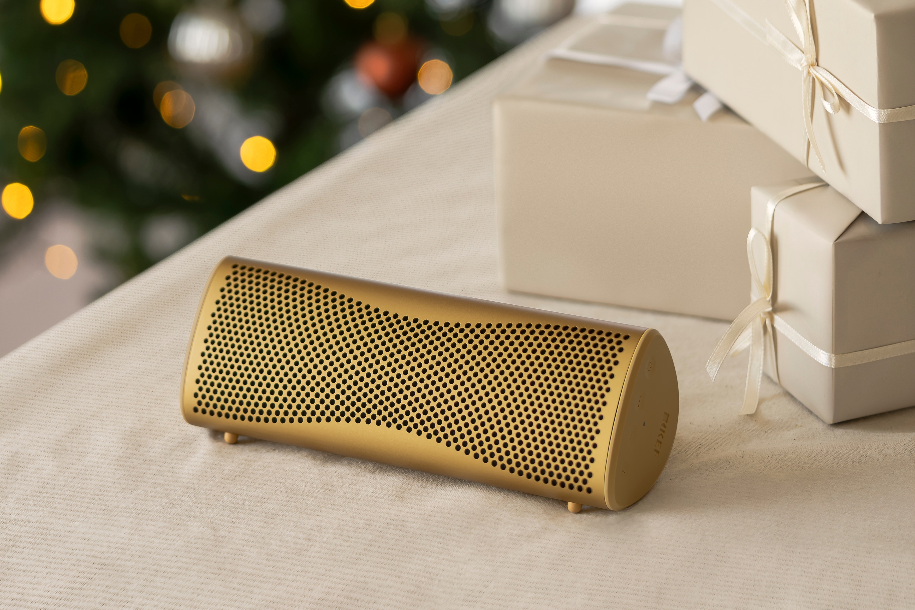 Gouden Bluetooth-speaker op een beige tafel naast ingepakte kerstcadeaus en een versierde kerstboom op de achtergrond.
