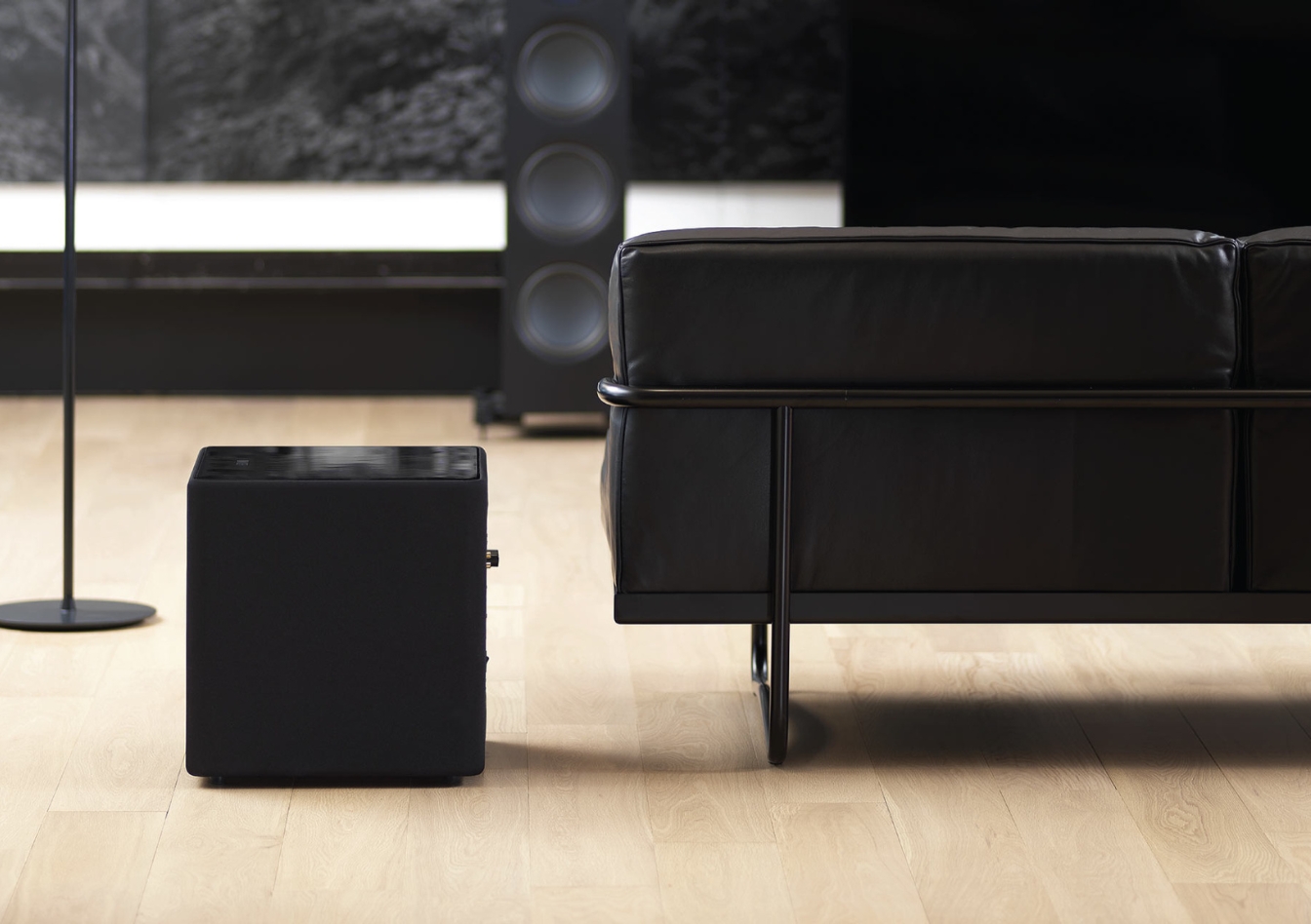 Compacte zwarte KEF subwoofer in een moderne woonkamer met leren bank en houten vloer