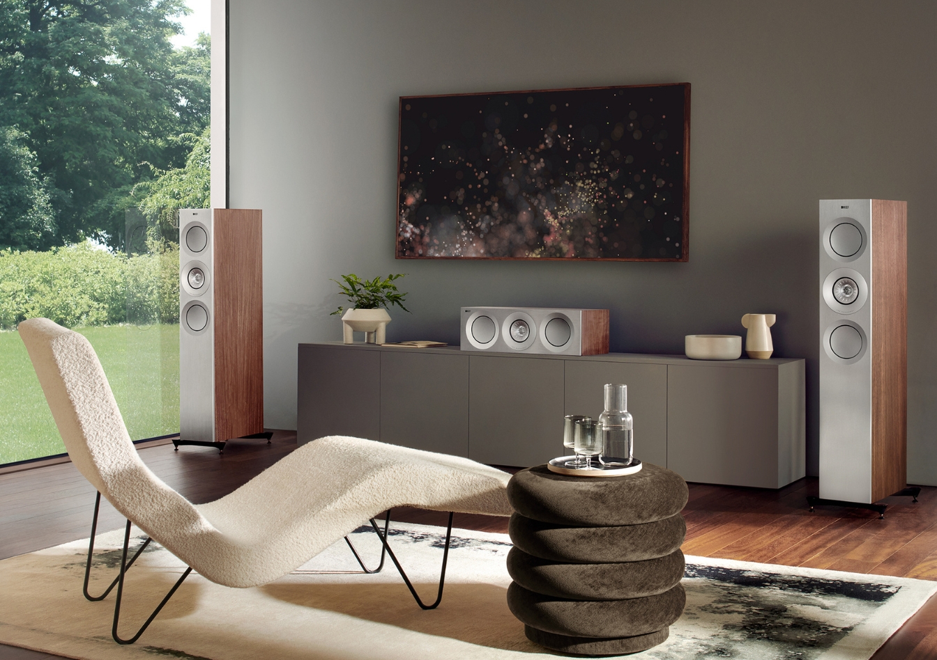 Elegante woonkamer met zilverkleurige vloerluidsprekers, bijpassende centerspeaker en houten accenten, opgesteld voor optimale audioplaatsing bij een groot raam met uitzicht op de tuin.