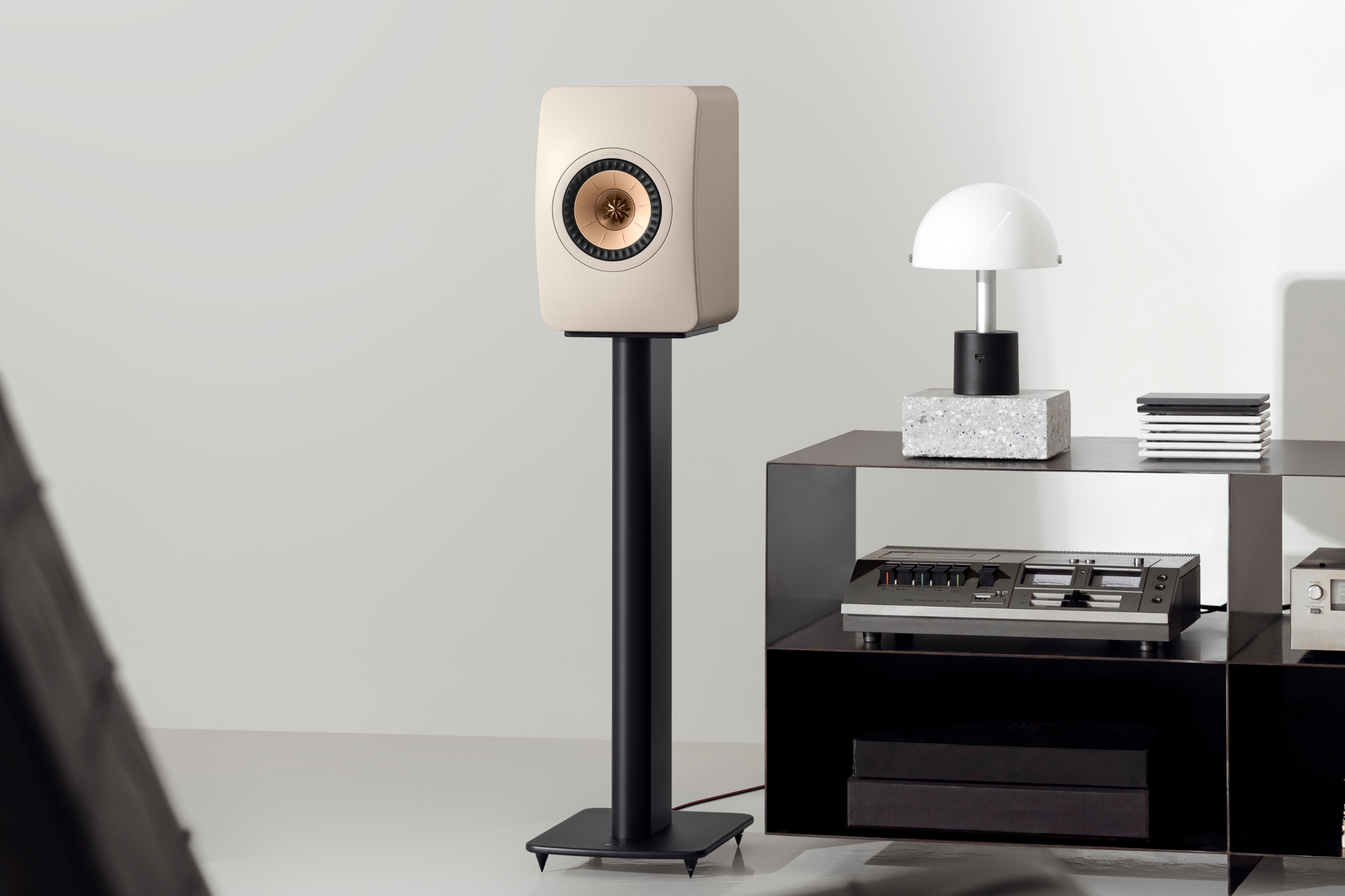 Moderne beige boekenplankspeaker op een zwarte vloerstandaard in een minimalistische woonkamer, naast een zwart rek met een retro hifi-installatie en een decoratieve lamp.