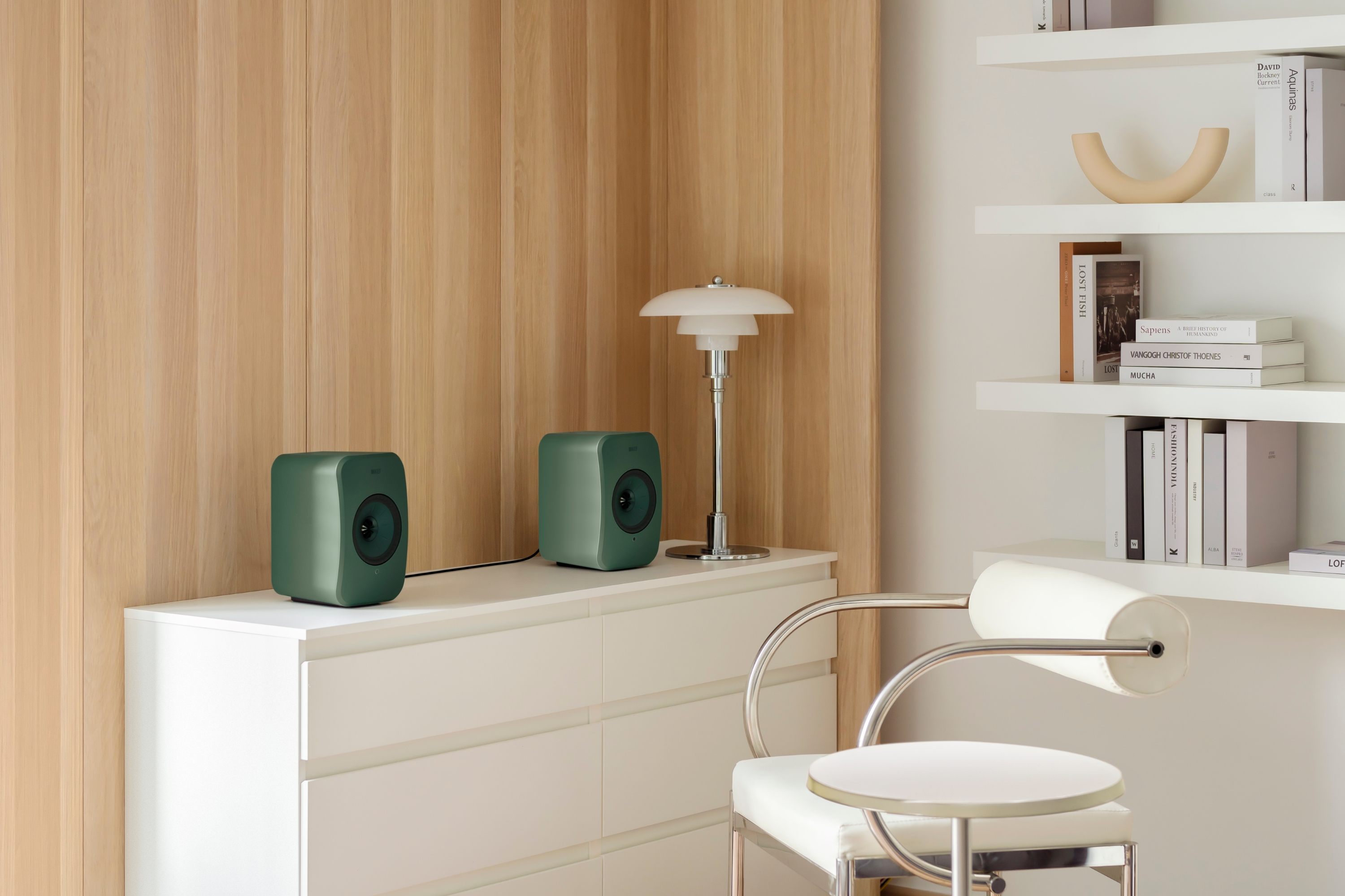 Een paar moderne groene boekenplankspeakers op een witte ladekast in een minimalistische kamer met houten lambrisering, witte planken en een moderne lamp.