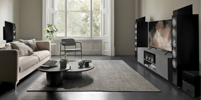 Moderne woonkamer met grote zwarte vloerluidsprekers en subwoofer, minimalistisch meubilair en een wandgemonteerde televisie, ontworpen om de ideale luidsprekerafmetingen voor thuisgebruik te demonstreren.