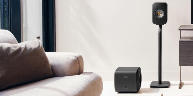 Compact KEF speaker on a stand with a matching subwoofer in a bright minimalist living room.