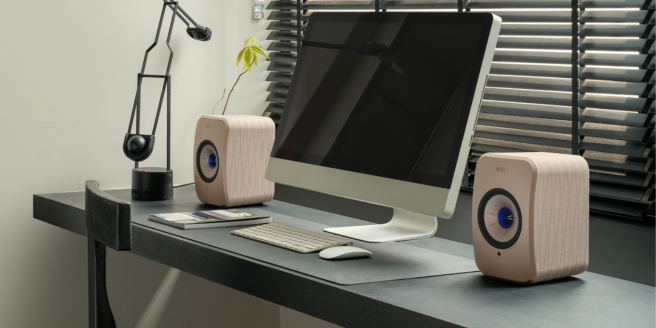 Two compact bookshelf speakers positioned on a modern desk beside an iMac computer in a home office.