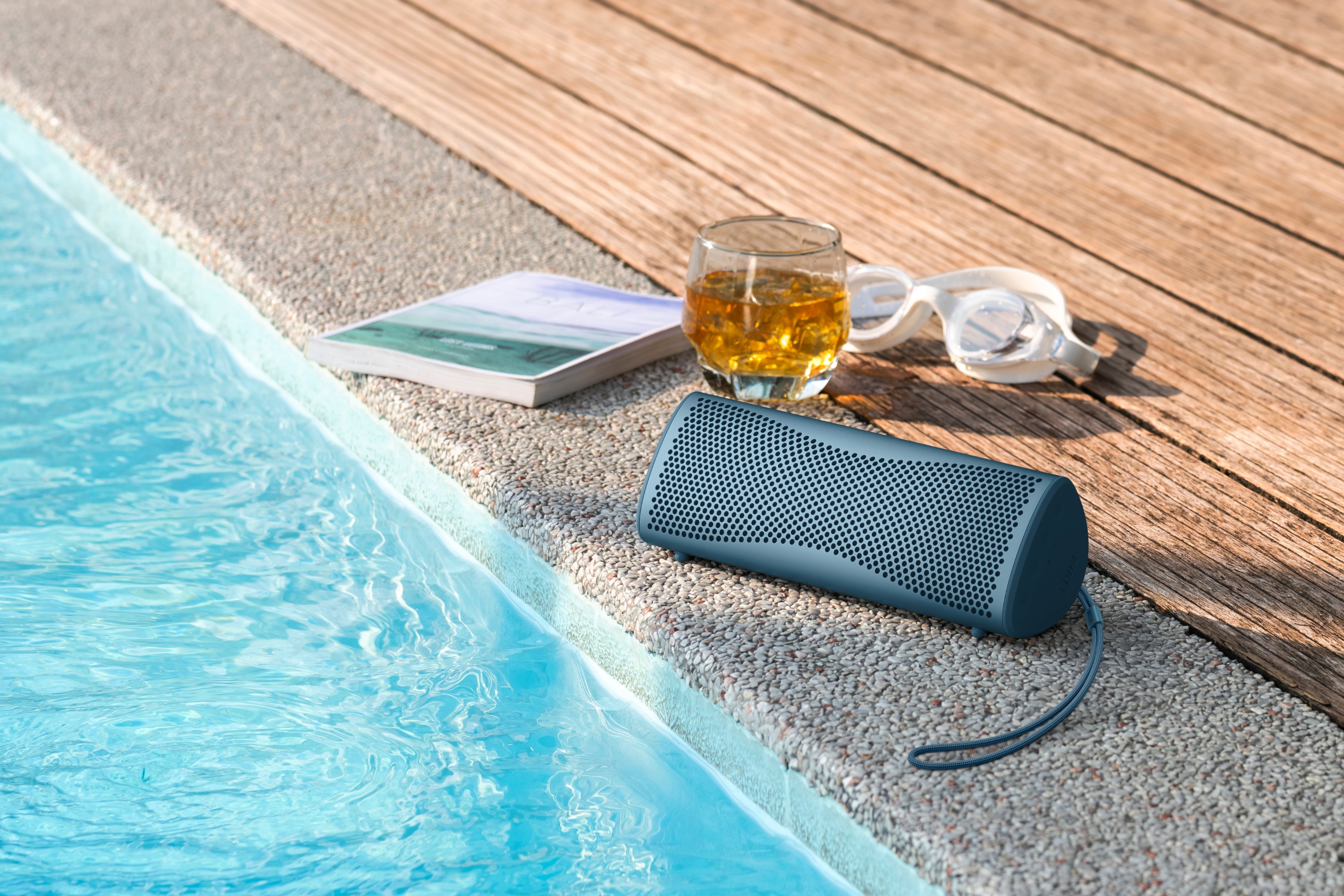 A portable Bluetooth speaker placed beside a swimming pool next to a book, drink and swimming goggles on a sunny day.