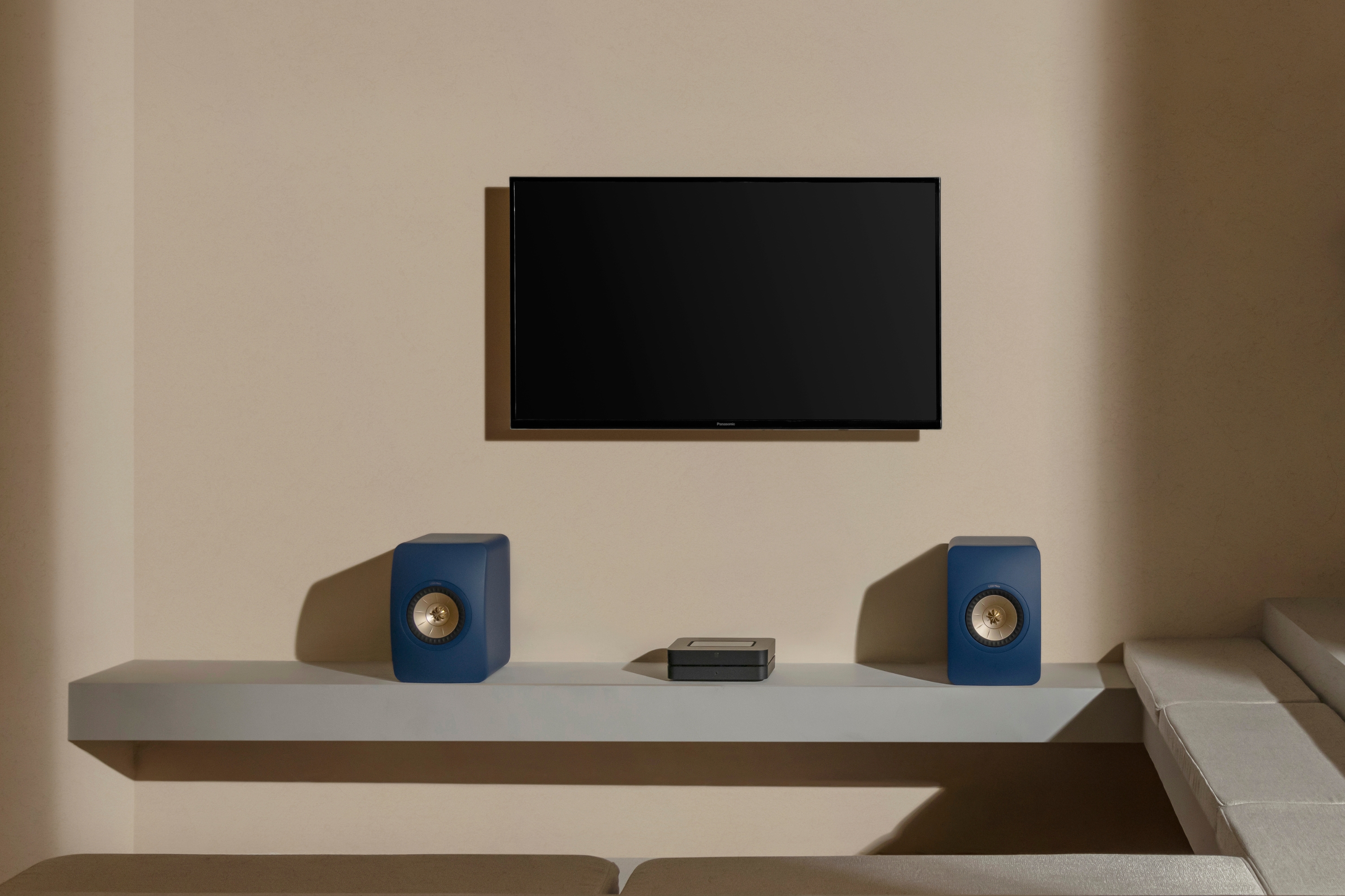 Installation home cinéma minimaliste avec une télévision murale, deux enceintes KEF LSX II bleues sur une étagère flottante, et une console multimédia centrale, le tout sur fond de mur beige.