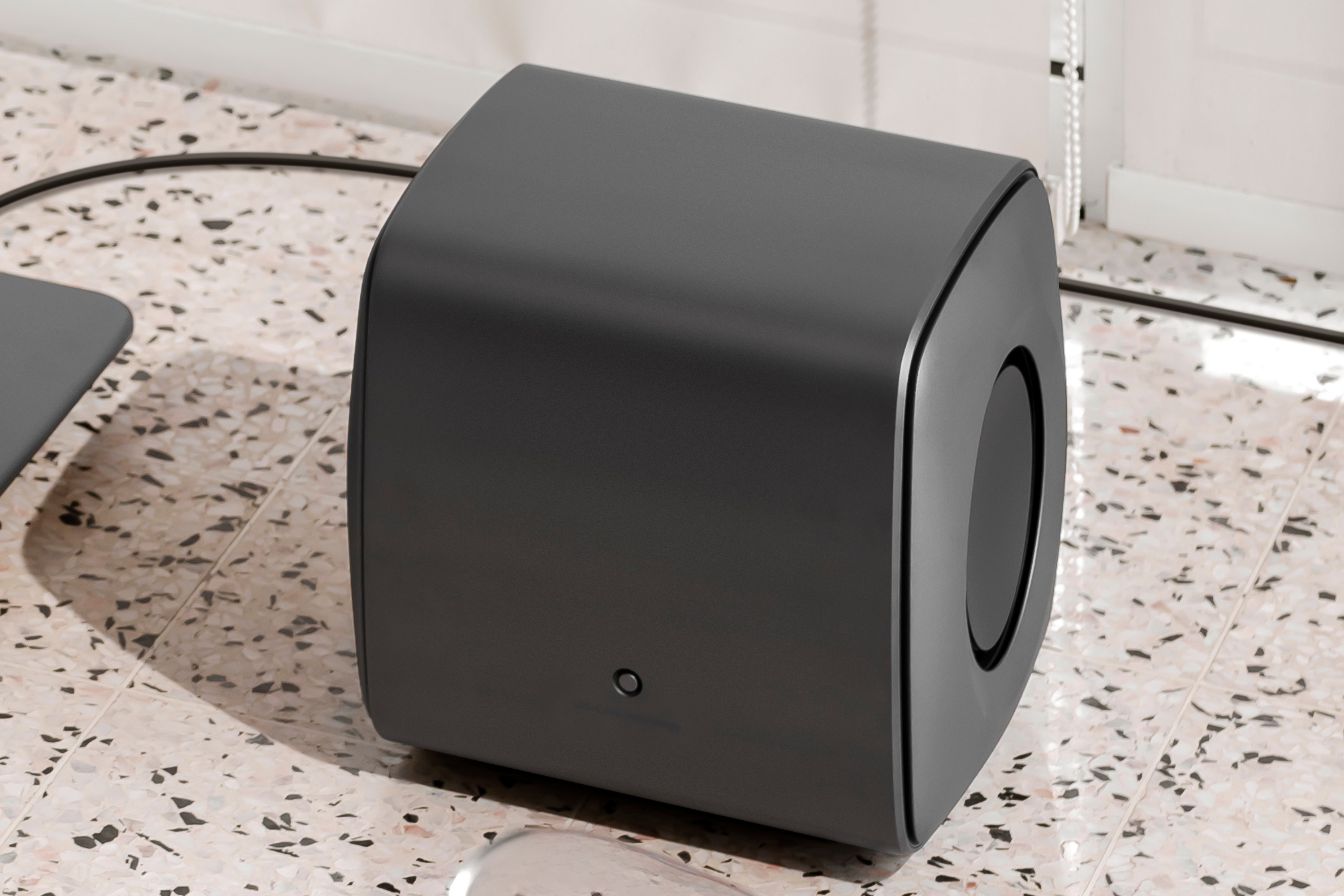 Caisson de basses KEF KC62 Uni-Core en finition noire mate, posé sur un sol en terrazzo, mettant en valeur son design compact et incurvé ainsi que ses haut-parleurs latéraux à annulation de force.