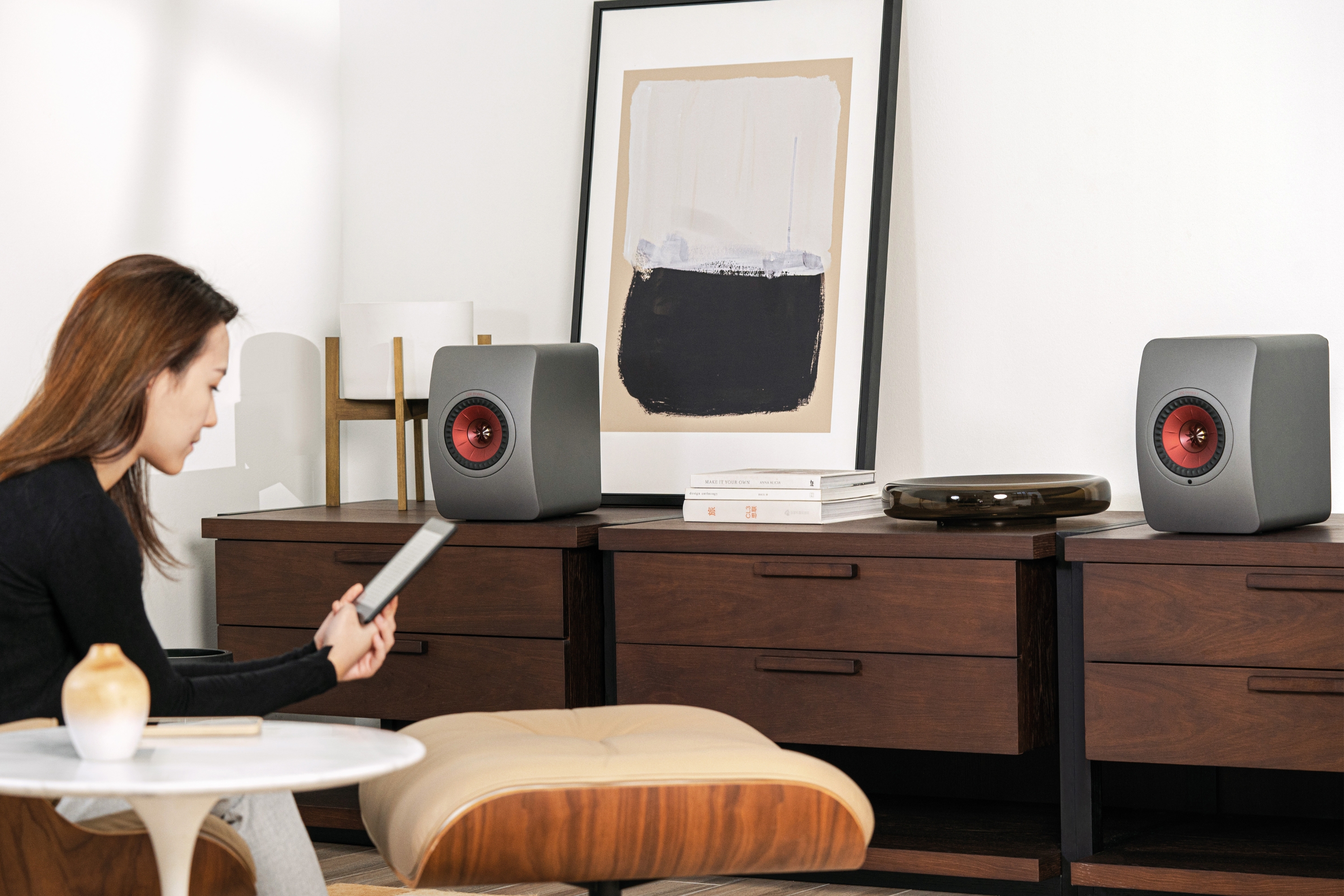 Eine Frau sitzt in einem modern eingerichteten Wohnzimmer und streamt Musik von einem Tablet auf ein Paar KEF LS50 Wireless II Lautsprecher in Carbon Black, die auf einem dunklen Holzsideboard mit minimalistischer Dekoration stehen.