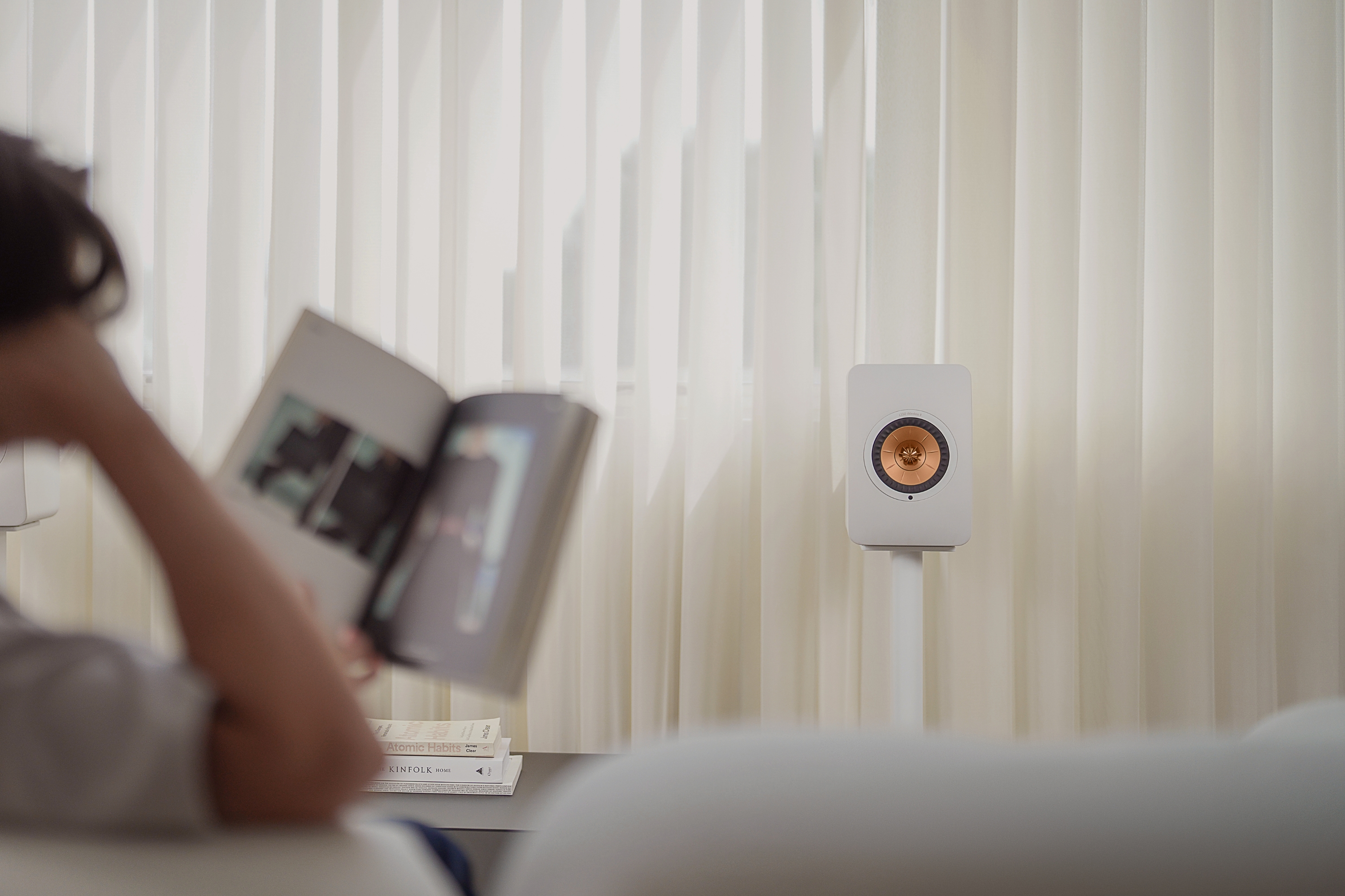 Eine Person entspannt sich mit einer Zeitschrift in einem sanft beleuchteten Raum, in dem ein weißer KEF LS50 Wireless II Lautsprecher auf einem Standfuß vor weißen Vertikaljalousien steht – für einen klaren, modernen Look.