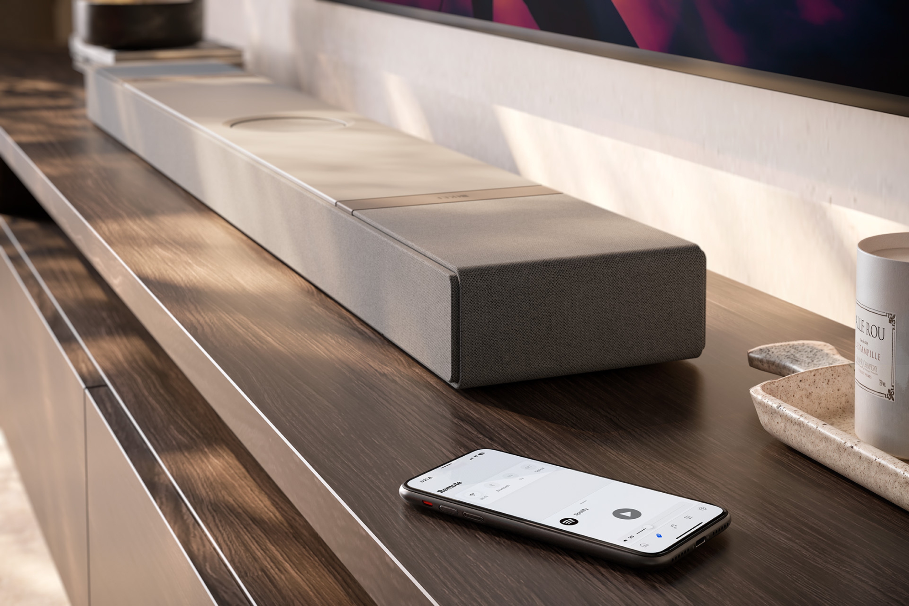 Close-up van een slanke KEF-soundbar op een houten mediameubel, met daarnaast een smartphone met een muziekapp – toont moeiteloze draadloze audiobediening.
