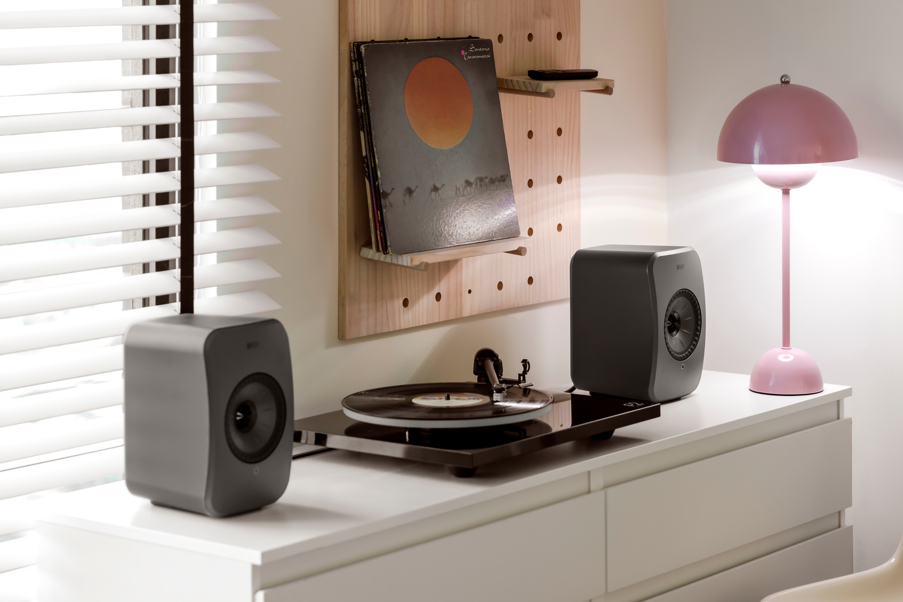 Paire d’enceintes de bibliothèque et une platine vinyle sur un buffet blanc, accompagnées d’une lampe rose et d’une présentation de disques vinyles dans un intérieur moderne.