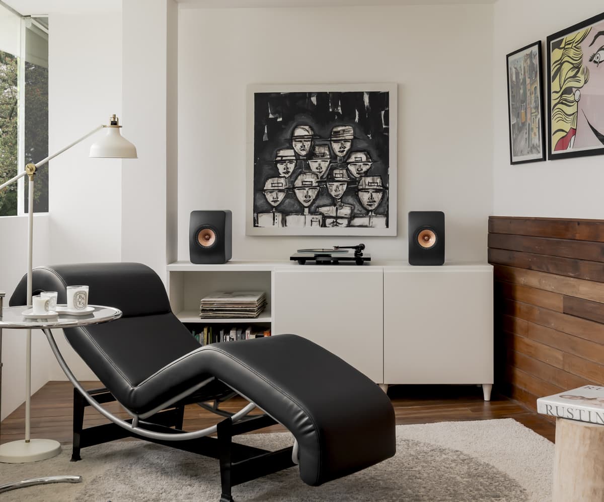Modern living room with black chaise lounge, turntable, and two LS50 Wireless II carbon black speakers on a white cabinet.