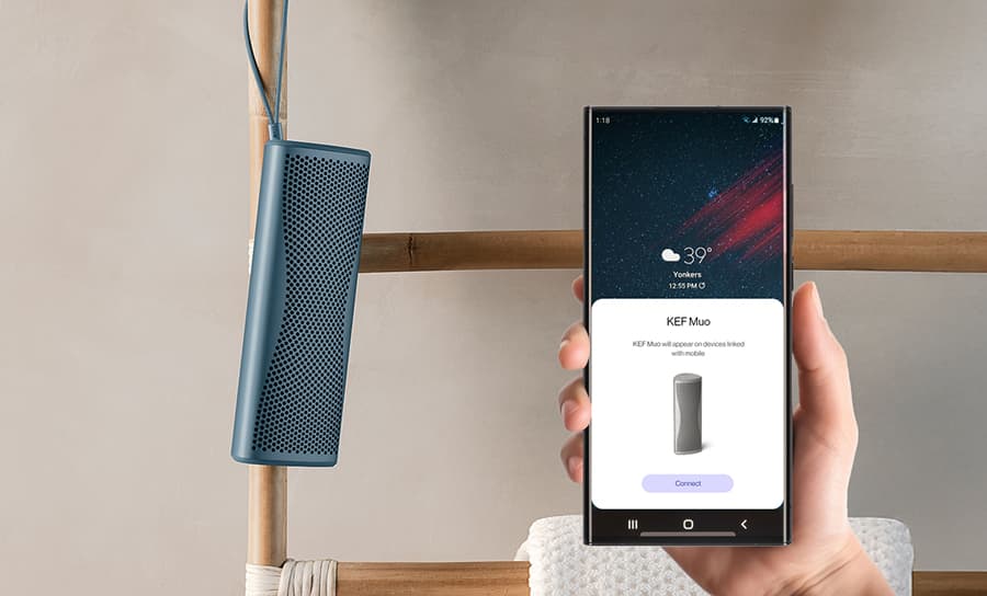 Enceinte Bluetooth Muo bleue suspendue par sa sangle de transport dans une salle de bains et contrôlée depuis un téléphone.